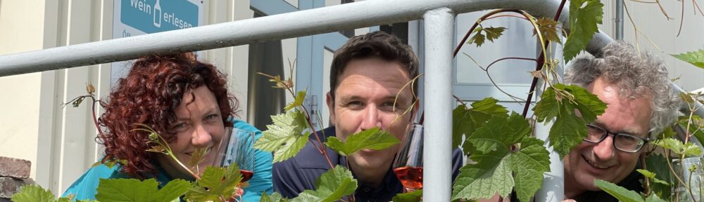 Das Bild zeigt das Team von weinerlesen, Anke Günther, Johannes Könitzer udn Dr. Olaf Nitzsche. Sie halten jeweils ein Weinglas in den Händen und schauen durch das Weinlauf eines Rebstocks in die Kamera. Im Hintergrund ist die Ladentür von wein-erlesen zu erkennen